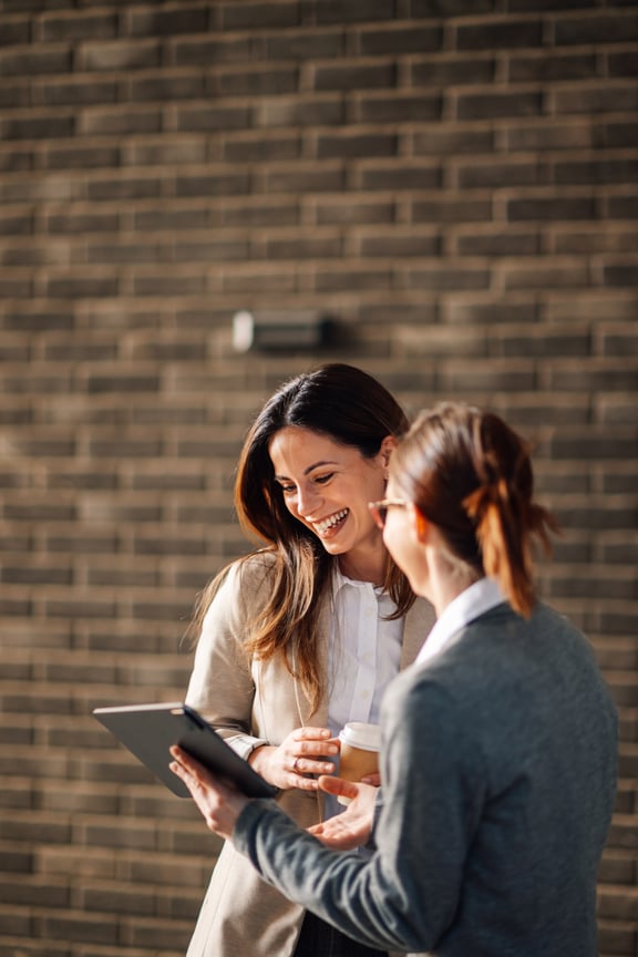 Zwei lachende Geschäftsfrauen, eine hält ein Tablet und zeigt der anderen etwas
